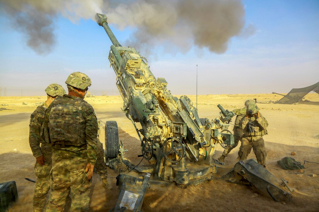 Smoke bursts out of a howitzer as soldiers fire it in desert terrain.