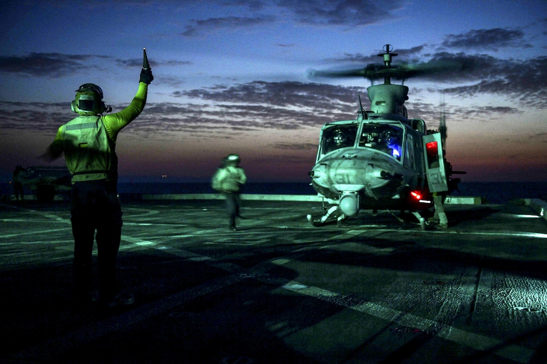 A sailor signals to a helicopter on a flight deck, lit up against a dim blue sky.