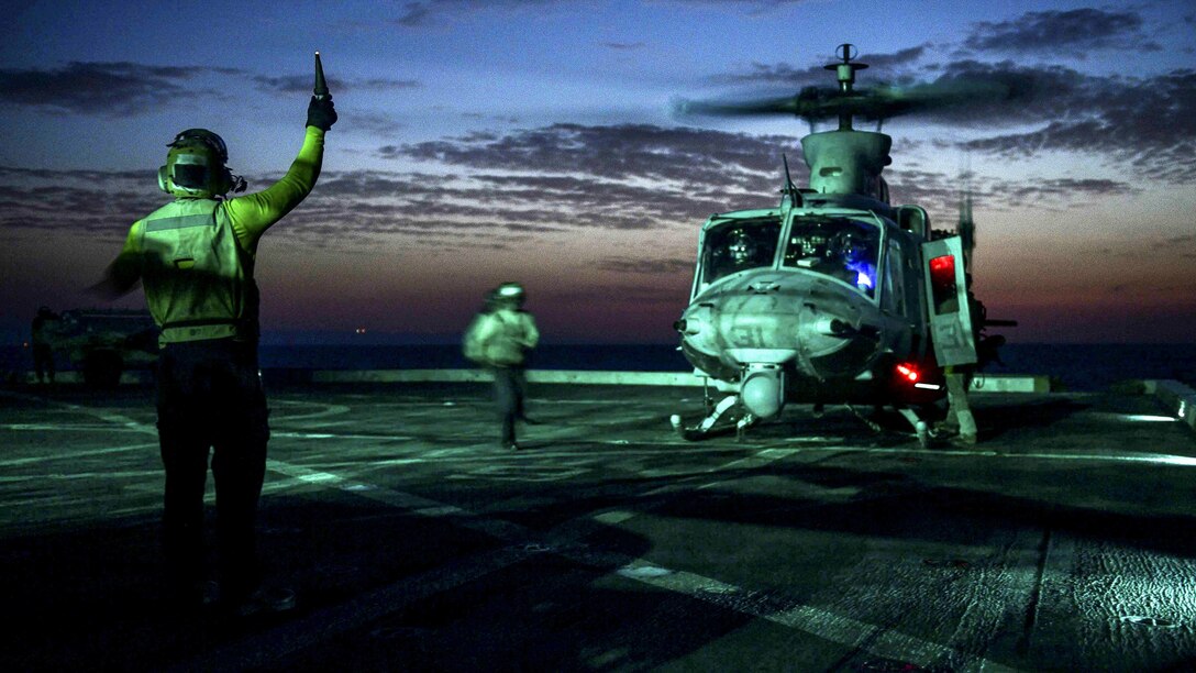 A sailor signals to a helicopter on a flight deck, lit up against a dim blue sky.
