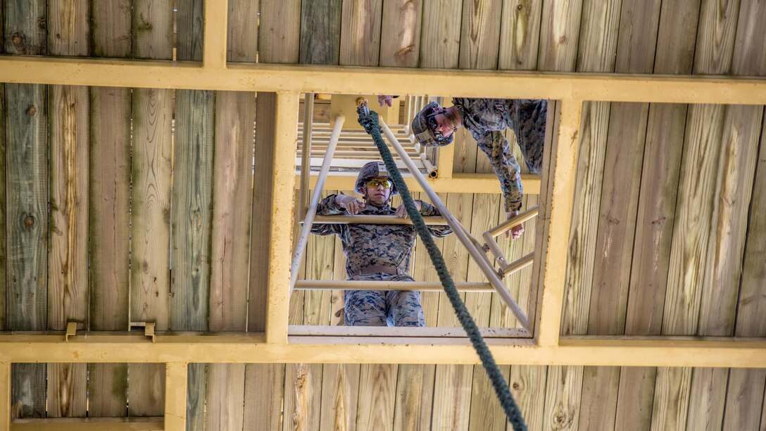 Two Marines look down through a square shaped opening in a wood floor that has a rope passing through it.