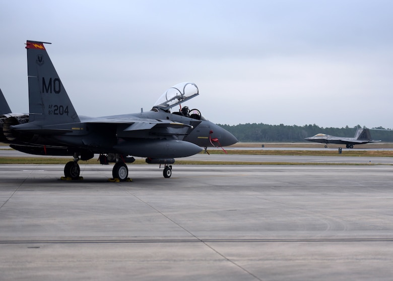 A U.S. Air Force F-15E Strike Eagle from the 389th Fighter Squadron, Mountain Home Air Force Base, Idaho, sits parked on the flightline as a 43rd Fighter squadron F-22 Raptor takes off in the distance at Tyndall Air Force Base, Fla., Nov. 9, 2017. Mountain Home was at Tyndall to participate Checkered Flag 18-1, a large-scale aerial exercise designed to integrate fourth and fifth-generation airframes while providing a platform for maintenance teams to be evaluated. (U.S. Air Force photo by Airman 1st Class Isaiah J. Soliz/Released)