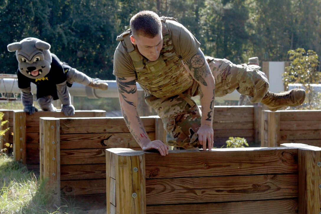 A soldier jumps over a hurdle, as a bulldog mascot him jumps over one behind him.