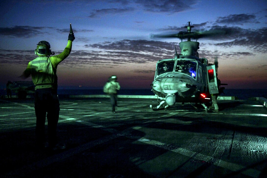 A sailor signals to a helicopter on a flight deck, lit up against a dim blue sky.