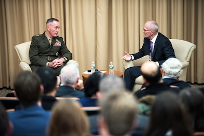 The chairman of the Joint Chiefs of Staff sits on a stage and talks to another person.
