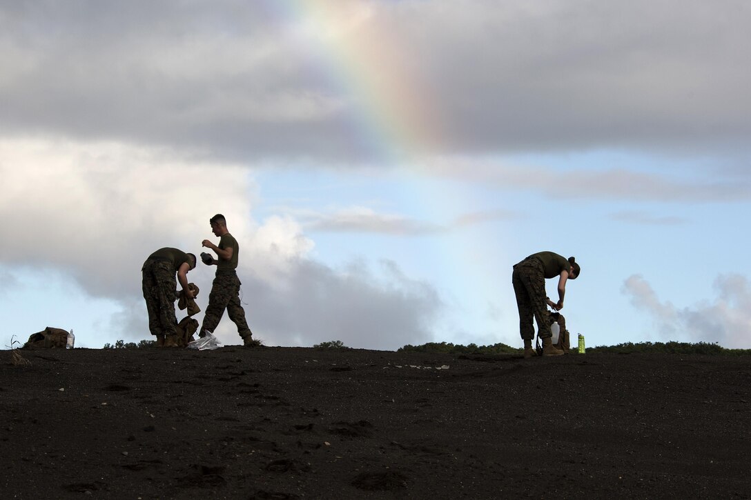 Two Marines bend over on sandy terrain and another walks, with a rainbow in the sky bend it.