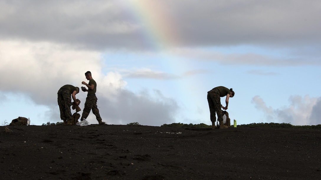 Two Marines bend over on sandy terrain and another walks, with a rainbow in the sky bend it.