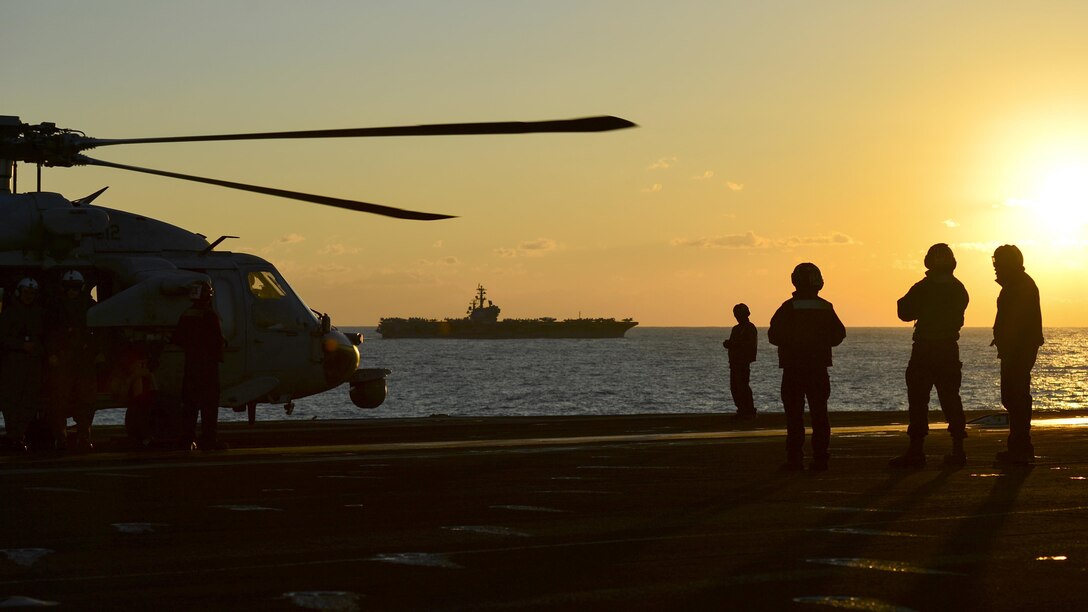 Four silhouetted photos stand on a ship's flight deck near a helicopter, against an orange sky.