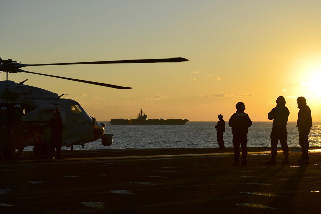 Four silhouetted photos stand on a ship's flight deck near a helicopter, against an orange sky.