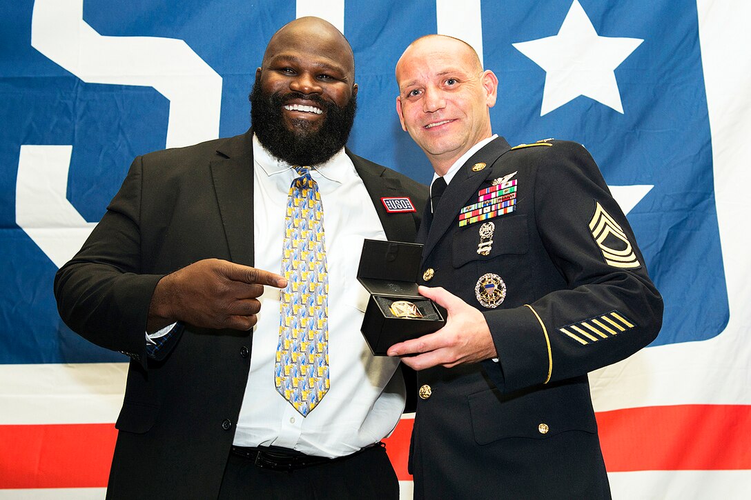 A master sergeant holds an award while standing next to a member of  World Wrestling Entertainment.