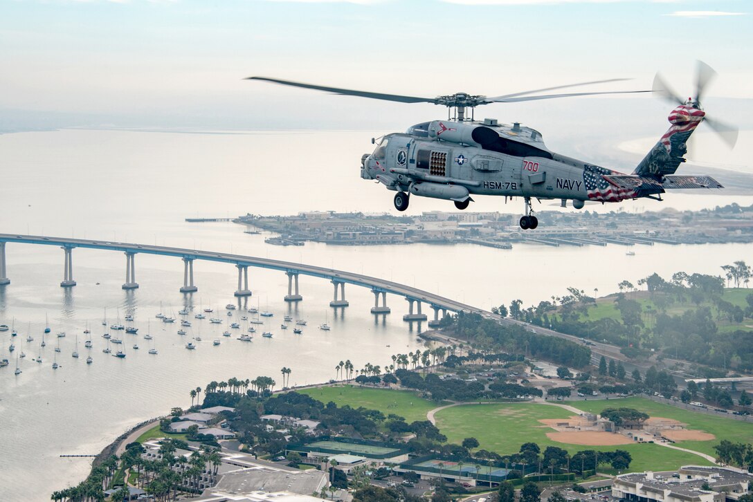 A helicopter flies over built-up coastline and water dotted with sailboats and spanned by a bridge.