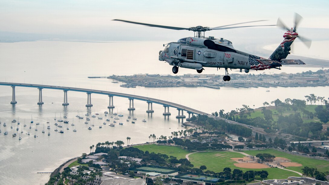 A helicopter flies over built-up coastline and water dotted with sailboats and spanned by a bridge.
