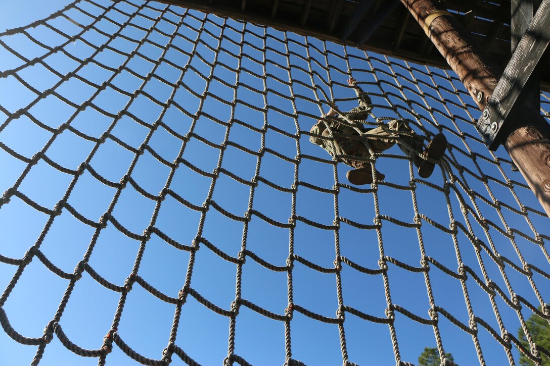 A soldier climbs a cargo net against a backdrop of blue sky.