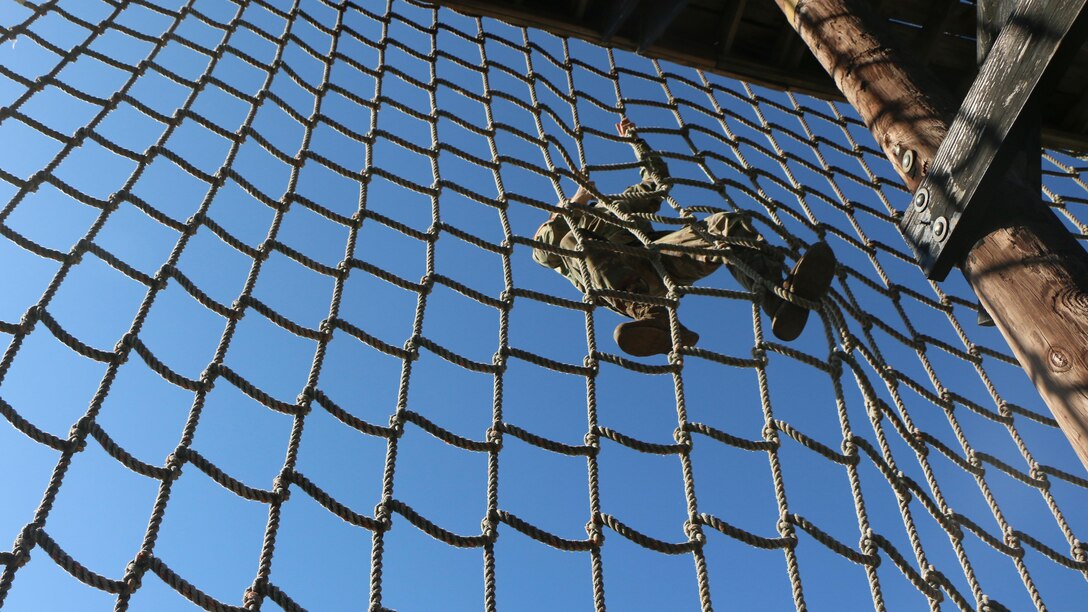 A soldier climbs a cargo net against a backdrop of blue sky.