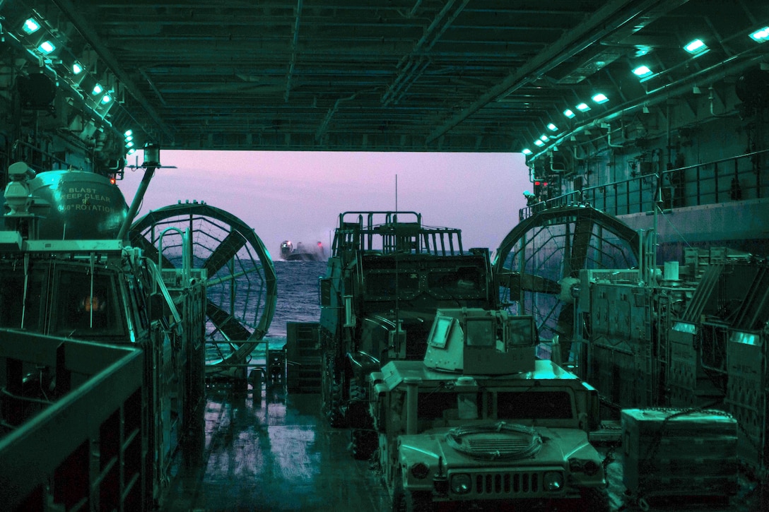 A landing craft approaches a well deck, filled with vehicles and illuminated in green.