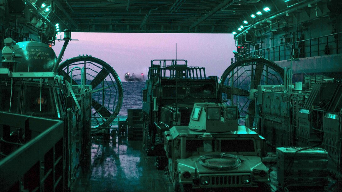 A landing craft approaches a well deck, filled with vehicles and illuminated in green.
