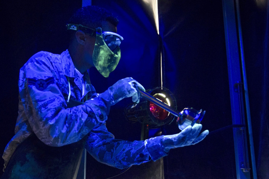 An airman wearing goggles and gloves inspects an item, against a deep blue background.