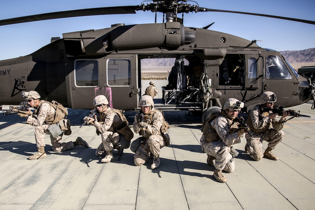 Marines kneel and point weapons while fanned out in front of a helicopter.