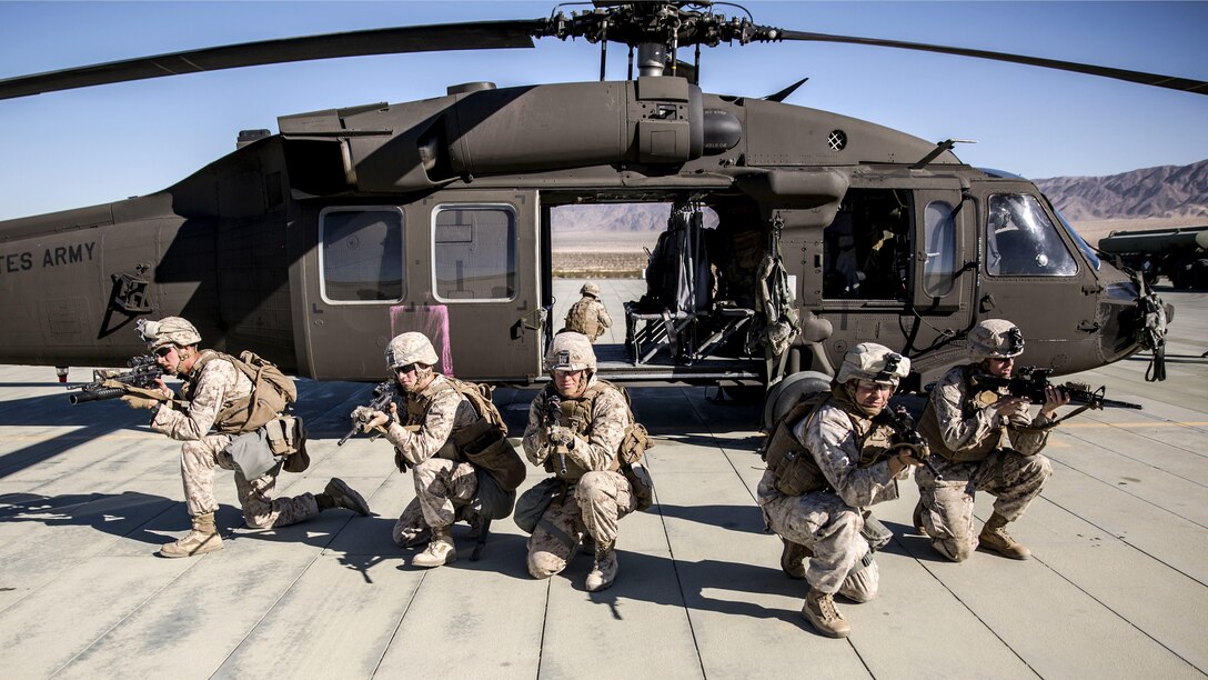 Marines kneel and point weapons while fanned out in front of a helicopter.