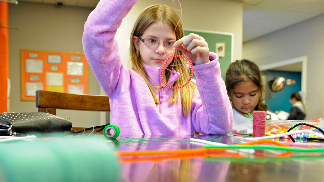 A child holds up a dreamcatcher and another works on her project during a crafts lesson.