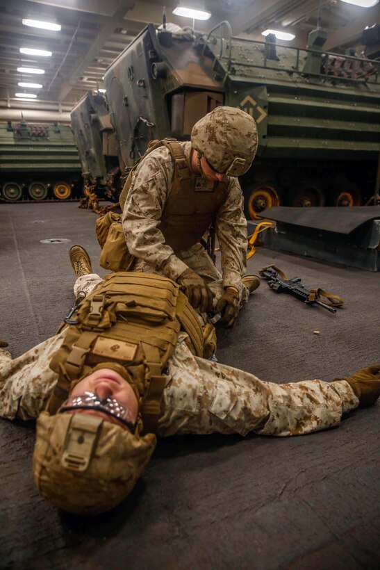 A Marine places a tourniquet to the leg of another Marine laying down.