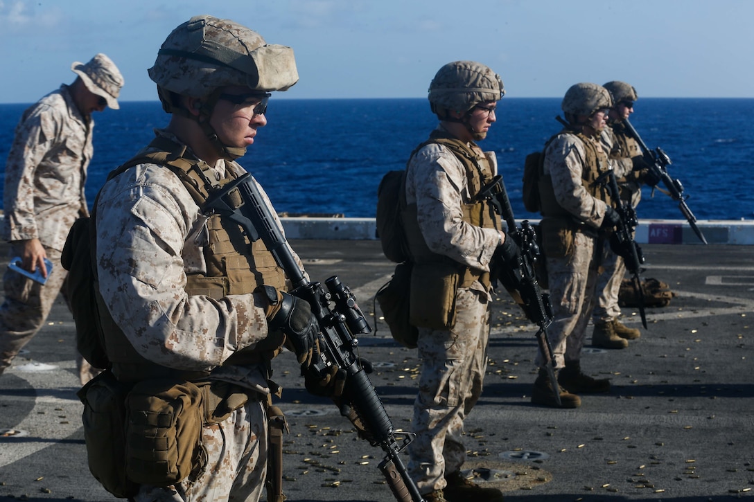 A line of Marines holds weapons while standing on the deck of a ship.