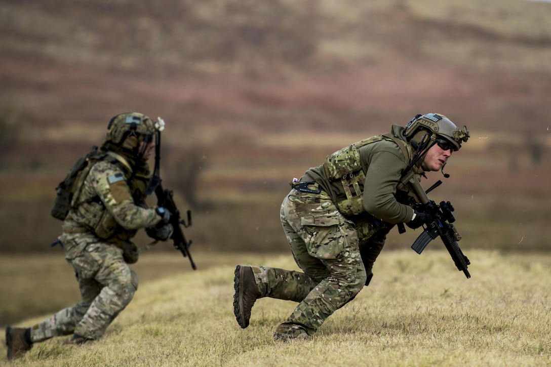 Two airmen wielding weapons lean forward while sprinting on slightly hilly terrain.
