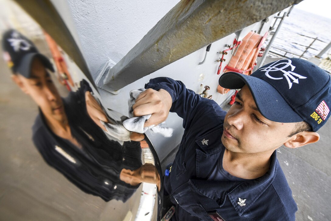 A sailor on a ship polishes a bell, which shows his reflection