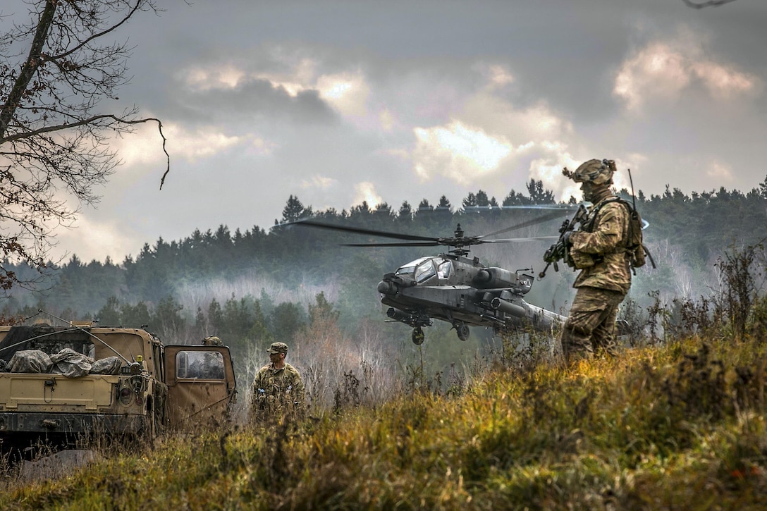 A helicopter lifts off in foggy weather in hilly terrain as soldiers stand nearby.