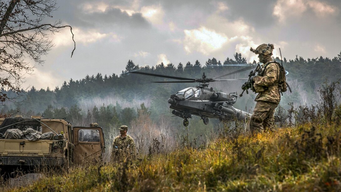 A helicopter lifts off in foggy weather in hilly terrain as soldiers stand nearby.