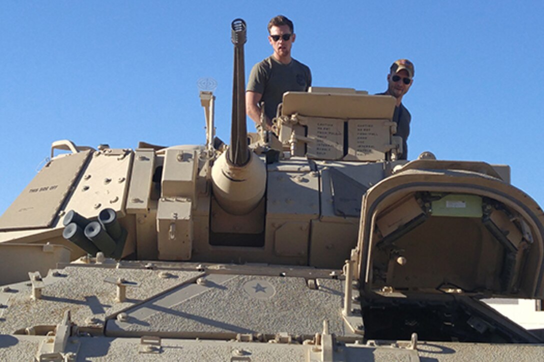 Two men stand on top of a military vehicle.