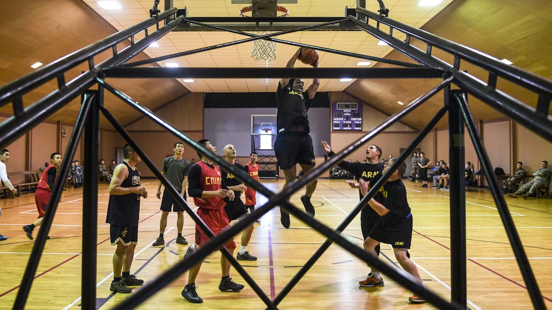 A service member jumps to take a shot during a basketball game, as others on the court watch.