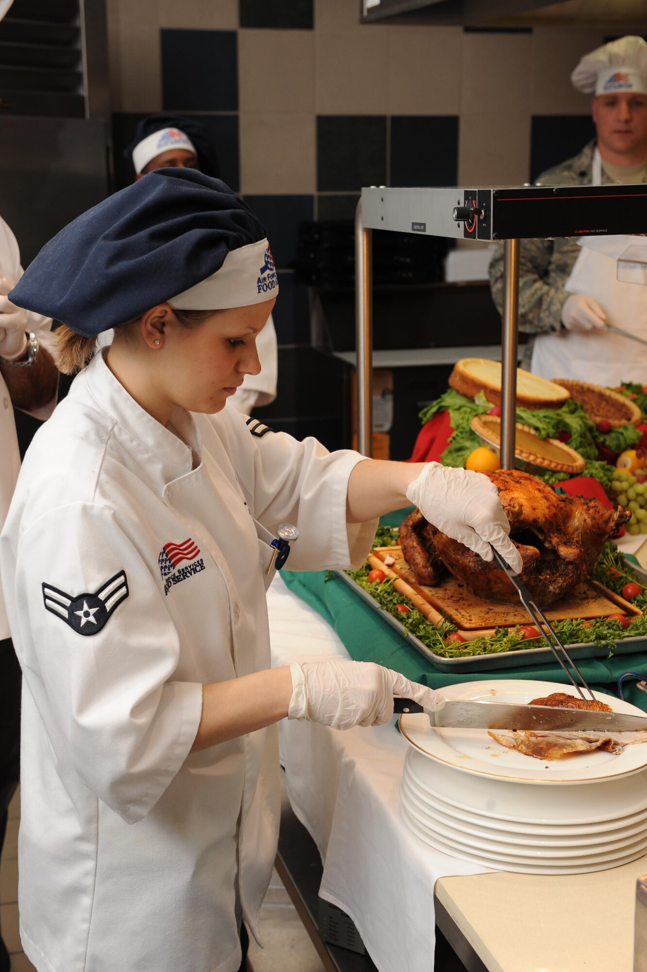 A female food service worker in white jacket and blue chefs hat, cuts turkey on a plate.