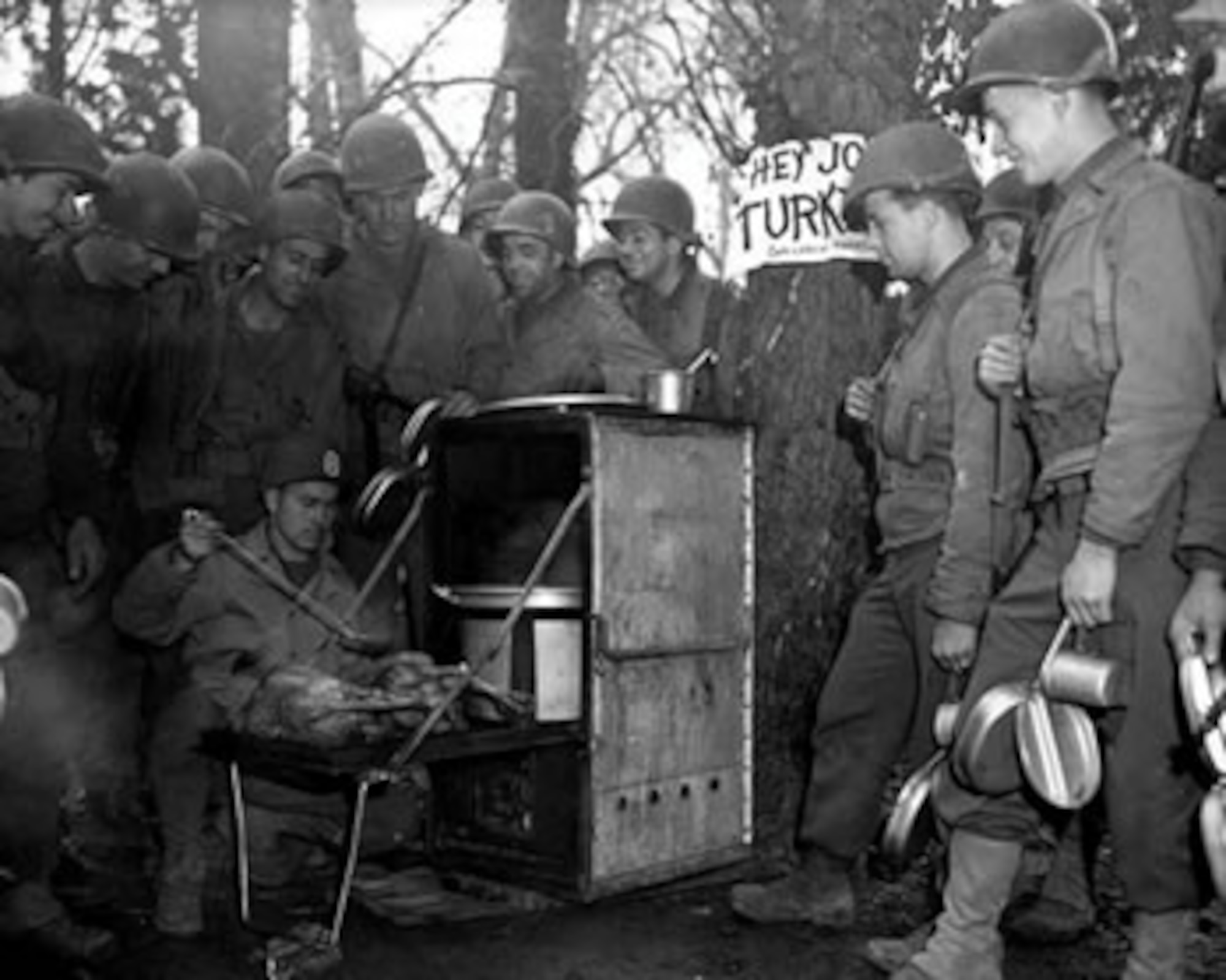 A male U.S. Army Sergeant prepares two Thanksgiving turkeys on a camp stove for his fellow male soldiers who surround him out in the woods.