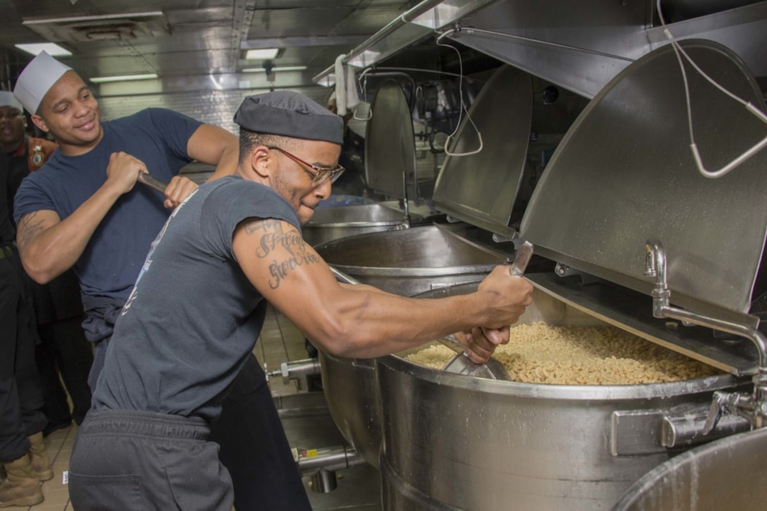 Two service members stir macaroni in huge vats on a ship.