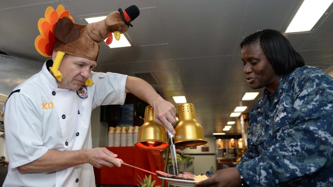 A man wearing a turkey hat serves meat to a female service member.