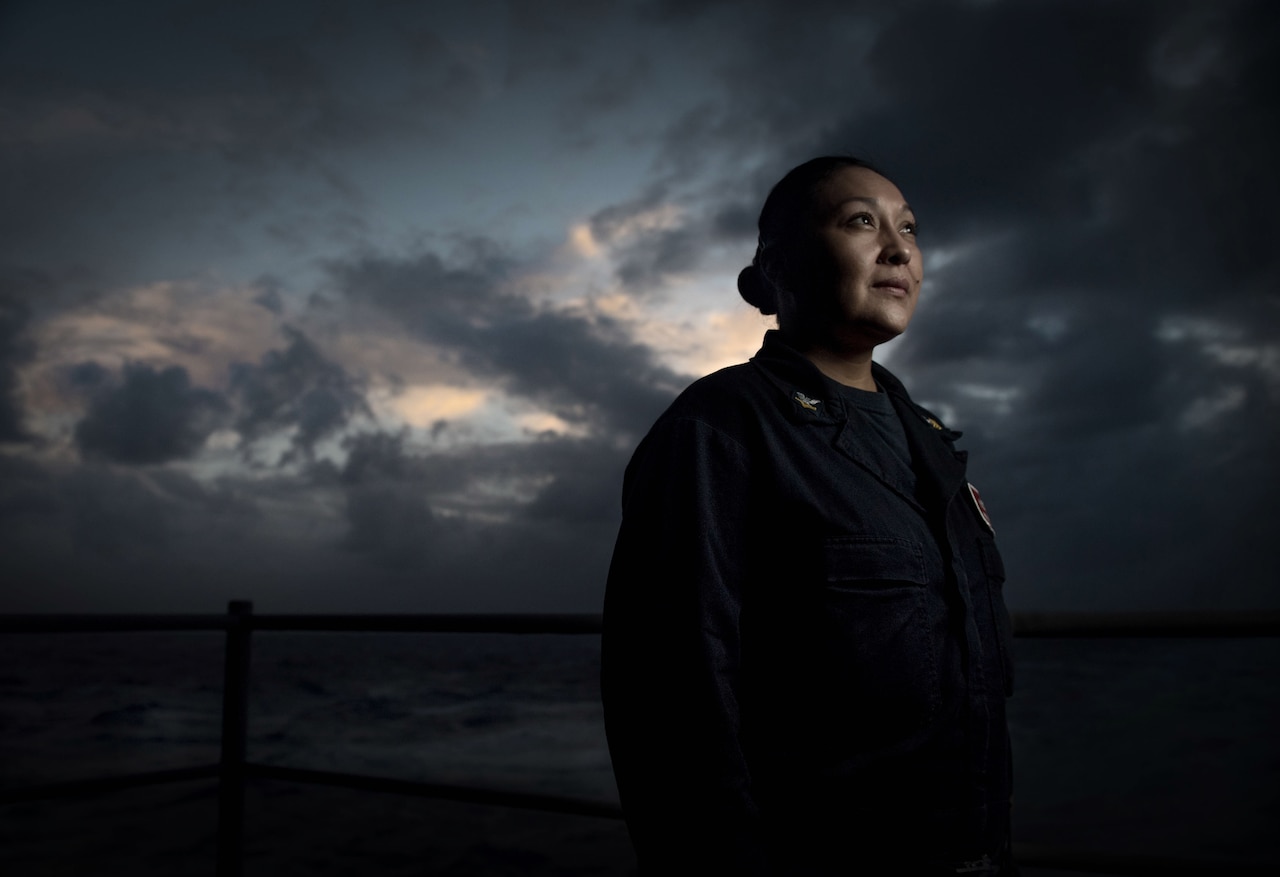 A sailor stands on the deck of a ship at night with dark clouds overhead.