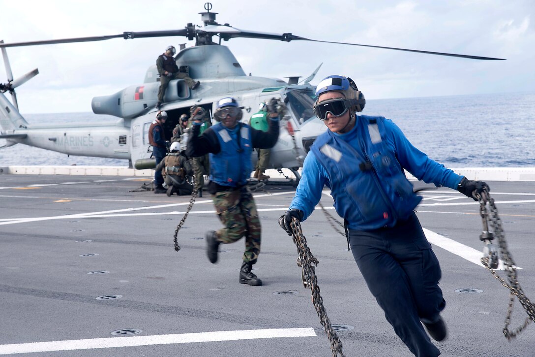 Two sailors carrying chains move away from a helicopter.