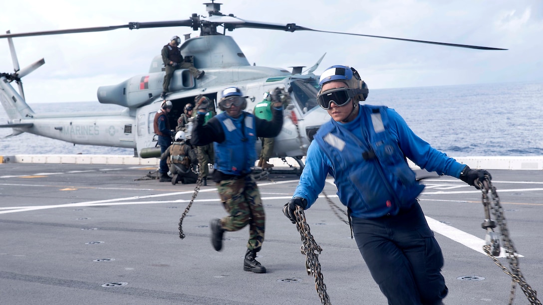 Two sailors carrying chains move away from a helicopter.
