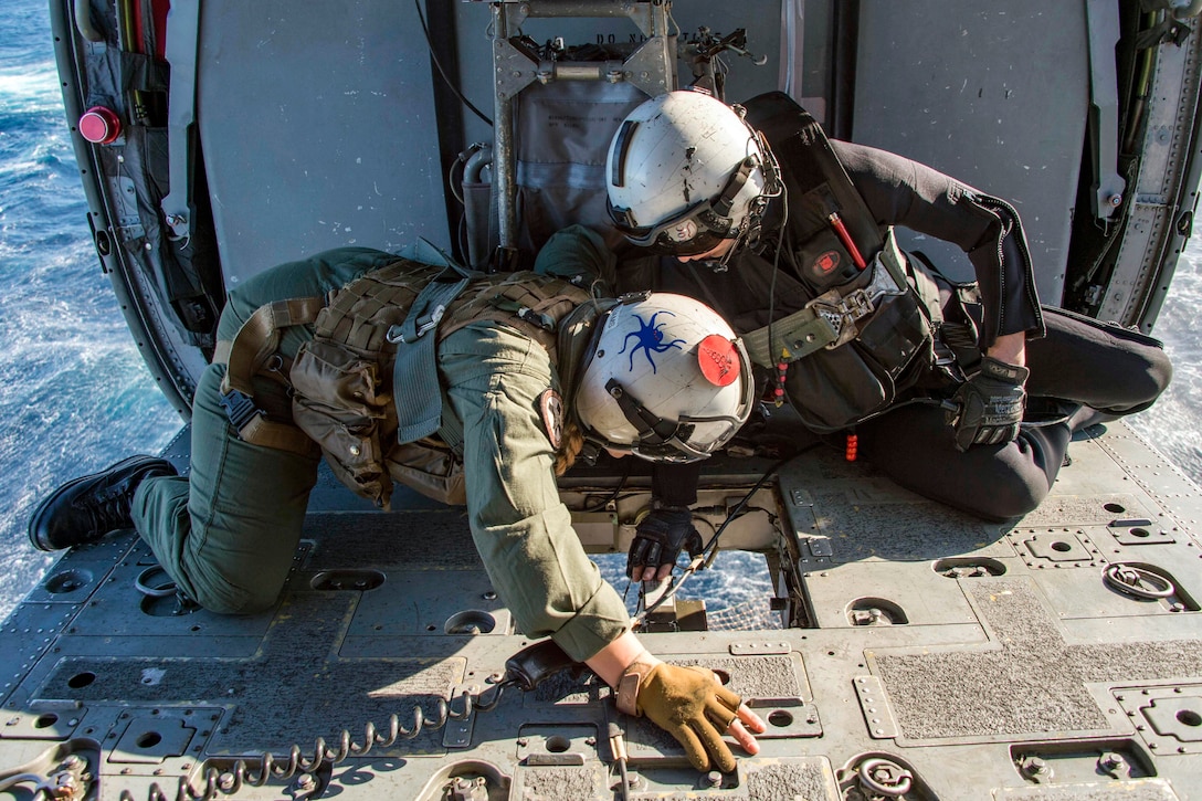 Two sailors look through a window in the bottom of a helicopter.