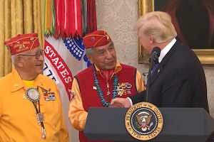 President Donald J. Trump shakes hands with two Navajo Code Talkers from behind a podium.