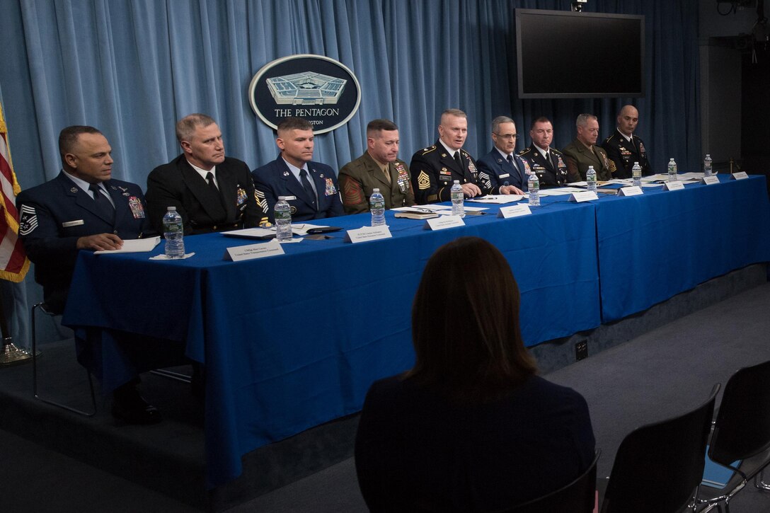 Military leaders sit at a long table and speak to reporters.