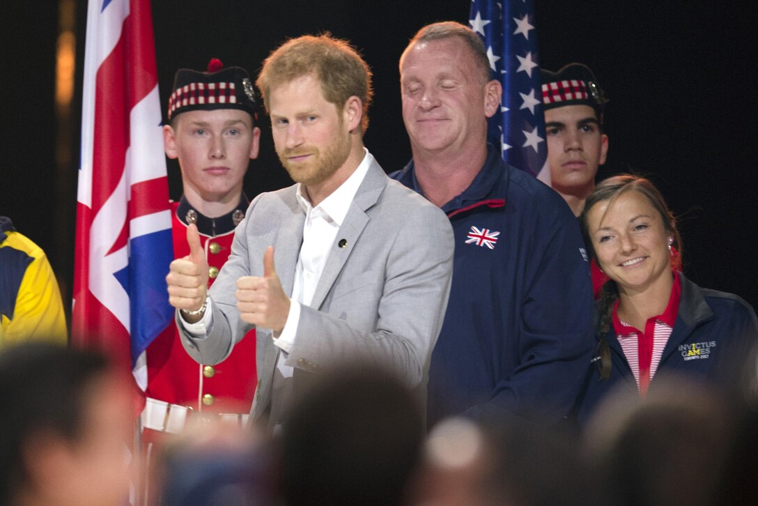 Prince Harry gives two thumbs up while surrounded by other people.