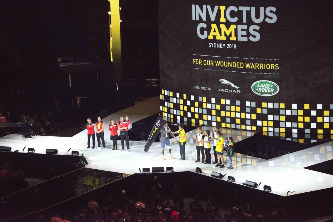 Two groups of people stand on a stage with a flag in the center.