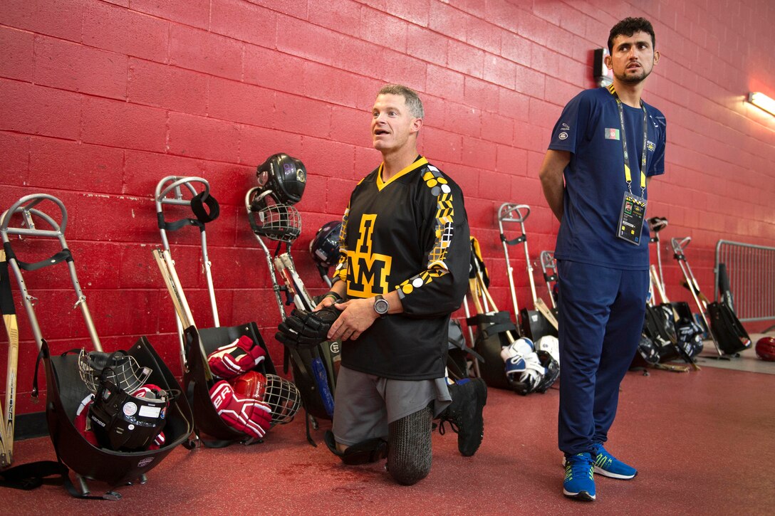 One person kneels and another stands next to sled hockey equipment.