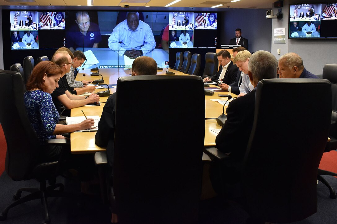 A group of people around a table look at teleconference screens.