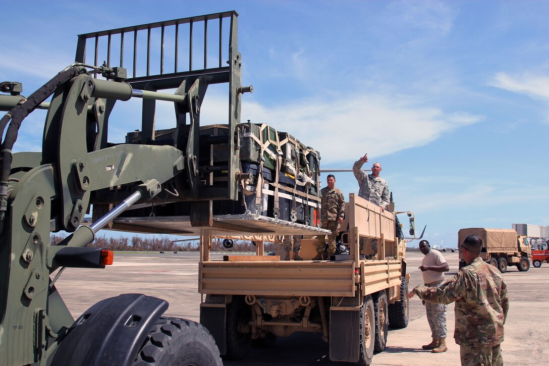 Service members direct and watch pallets of supplies being loaded into a military vehicle.
