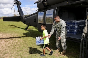 A soldier watches a child carrying a case of water away from a helicopter.