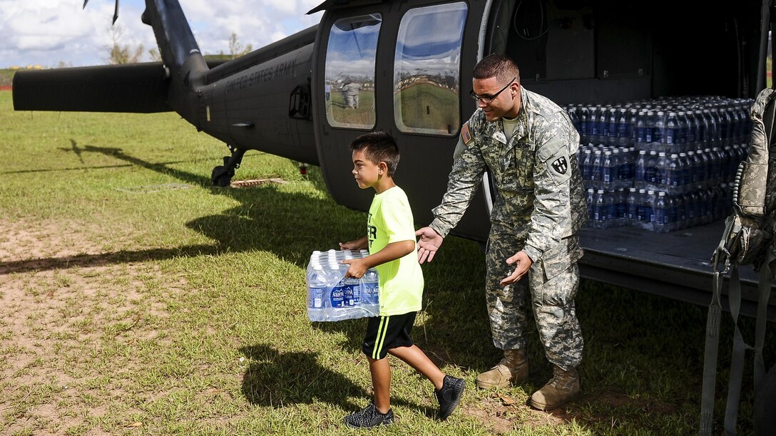 A soldier watches a child carrying a case of water away from a helicopter.