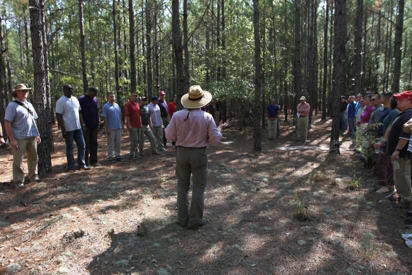 Two groups of people stand facing eachother in the woods while a guide speaks.