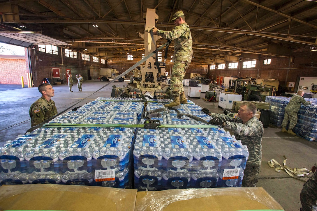 Guardsmen tighten and secure cargo straps around a large pallet of water before loading it on a heavy expanded mobility tactical truck.